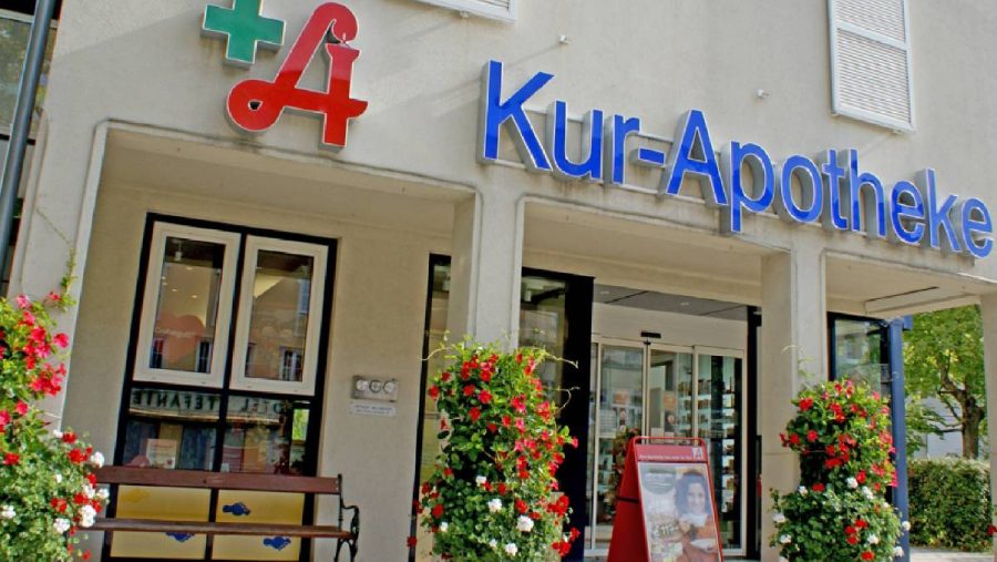 Eingang einer deutschen Apotheke mit Schaufenstern und dekorativen Blumenarrangements.