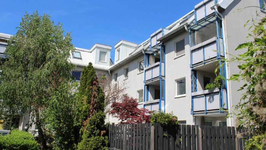 Mehrfamilienhaus mit blauen Balkonen und Garten im Vordergrund unter blauem Himmel.