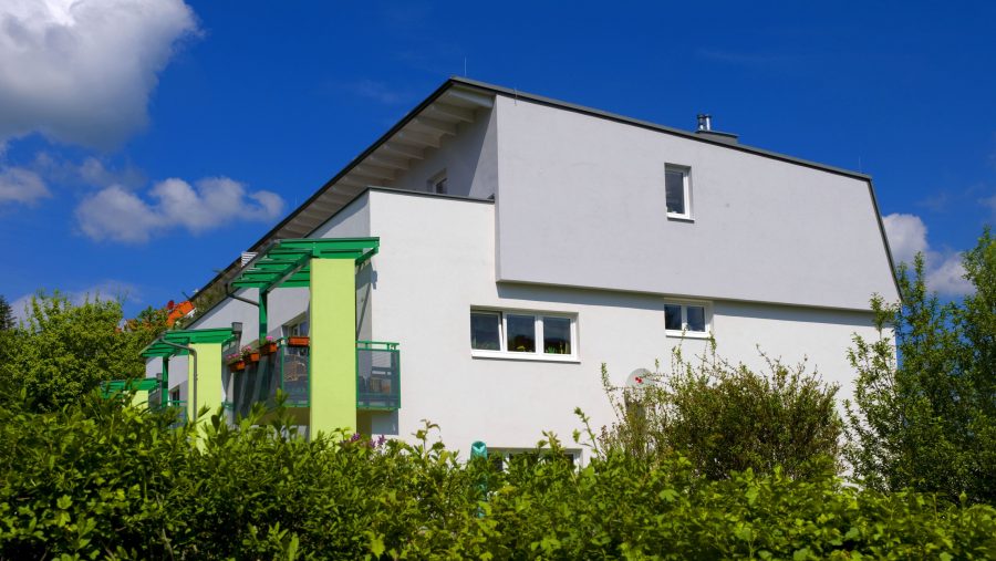 Modernes weißes Haus mit Balkon und grünem Geländer vor blauem Himmel und Wolken.