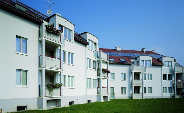 Mehrfamilienhaus mit Balkonen, Blumen und Solarpanelen bei klarem Himmel.