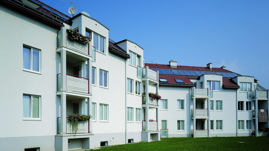 Mehrfamilienhaus mit Balkonen, Blumen und Solarpanelen bei klarem Himmel.