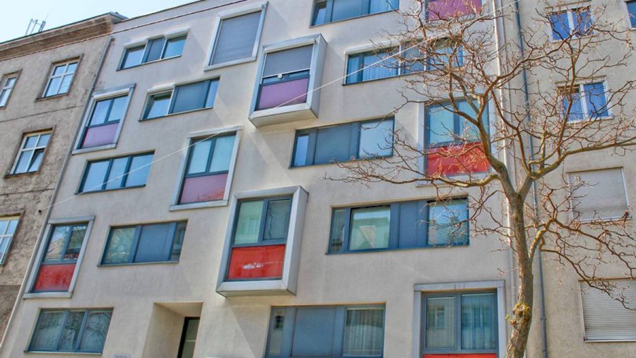 Moderne Wohnhausfassade mit farbigen Akzenten und kahlem Baum im Vordergrund.