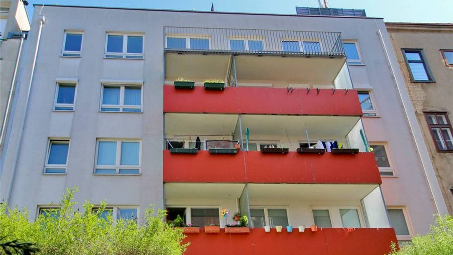 Mehrstöckiges Wohnhaus mit Balkonen und roten Brüstungen, umgeben von Pflanzen.