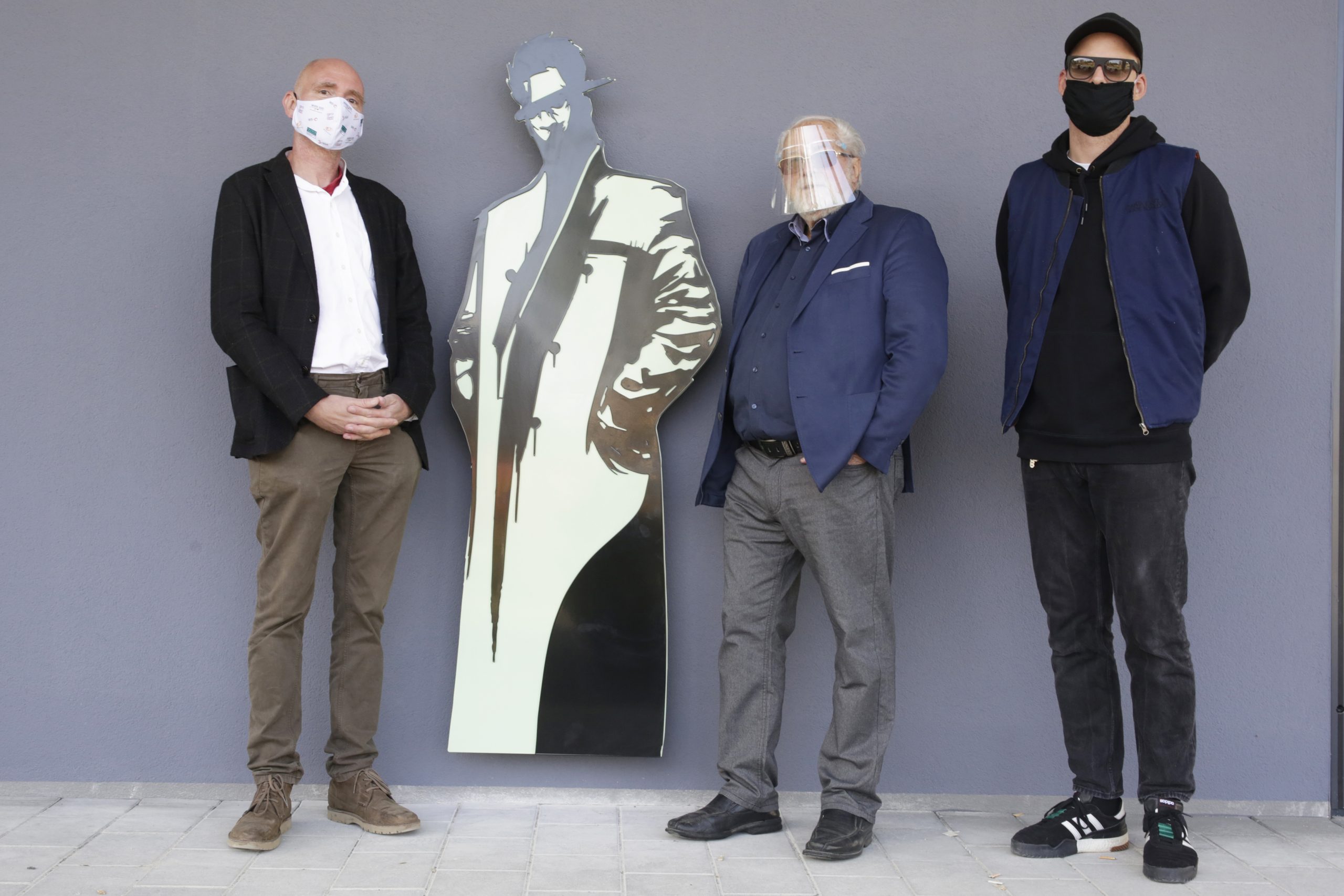 E0A1378 Rania Moslam Drei Personen mit Masken stehen neben einer stylisierten Silhouette vor einer grauen Wand.