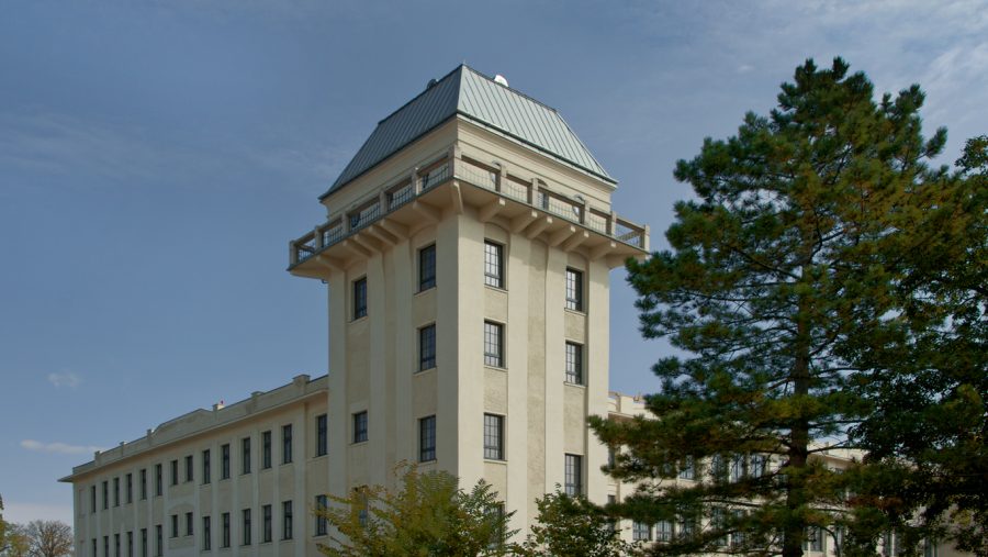 Großes, weißes Gebäude mit grauem Dach und vielen Fenstern, umgeben von Bäumen und blauem Himmel im Hintergrund.