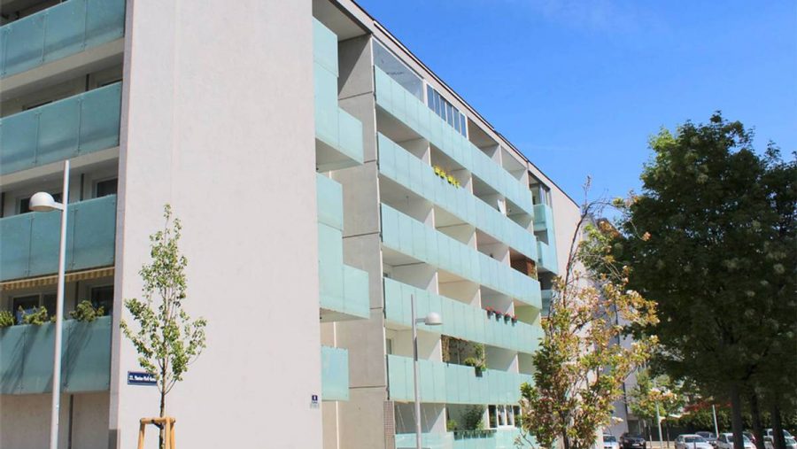 Mehrstöckiges Wohngebäude mit Balkonen und Bäumen im Vordergrund bei klarem Himmel.