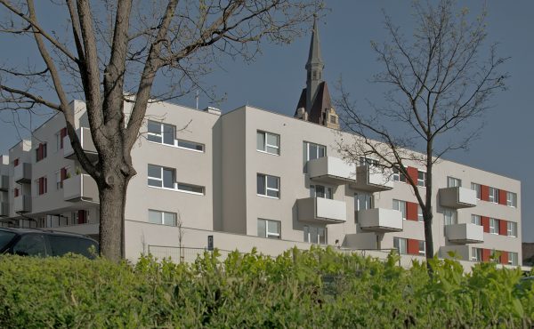 Modernes Wohngebäude mit Balkonen neben einem Kirchturm, umgeben von Bäumen und Sträuchern.