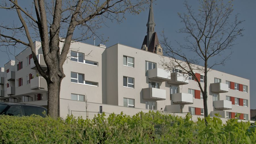Modernes Wohngebäude mit Balkonen neben einem Kirchturm, umgeben von Bäumen und Sträuchern.