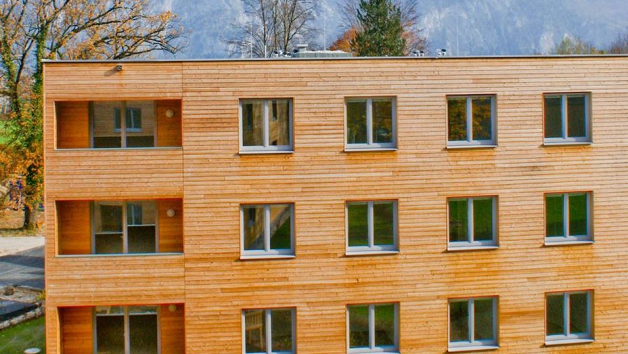 Modernes Holzgebäude mit mehreren Fenstern, vor einer bergigen Landschaft.