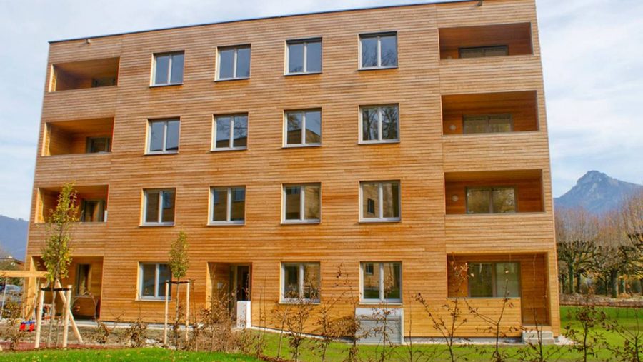 Mehrfamilienhaus aus Holz mit Balkonen, umgeben von Gras und Bäumen vor einem Bergpanorama.