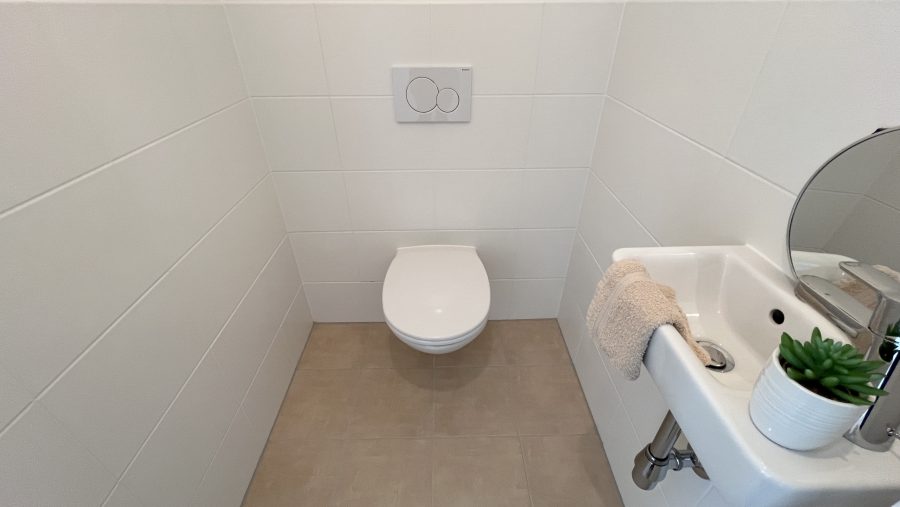 Kleines Badezimmer mit Toilette, Waschbecken und Pflanze auf heller Fliesenwand.