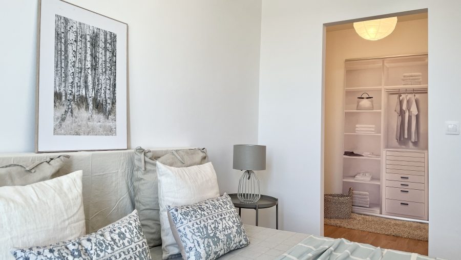 Minimalistisches Schlafzimmer mit Kunstwerk an der Wand, Tischlampe und offenem Kleiderschrank im Hintergrund.