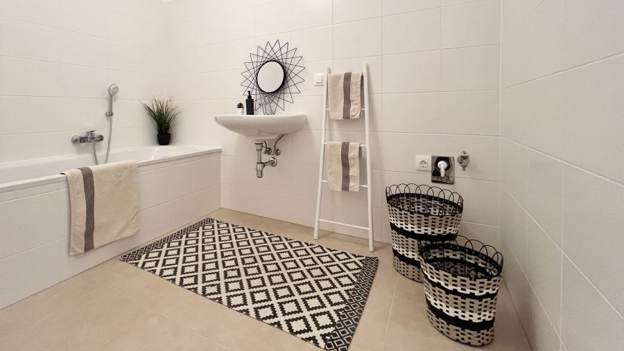 Modernes Badezimmer mit weißer Badewanne, Waschbecken, Handtüchern und dekorativem Teppich auf Fliesenboden.