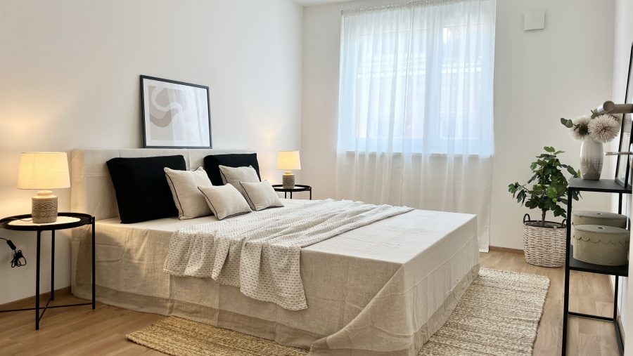 Modernes Schlafzimmer mit Doppelbett, neutralen Farben und Pflanzen, hell beleuchtet durch ein Fenster mit Vorhängen.