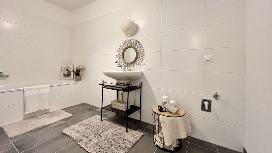 Modernes Badezimmer mit weißer Fliesenwand, Waschbecken, Spiegel und grauem Fliesenboden.