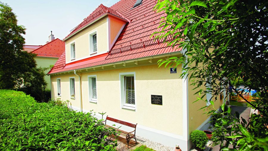 Gelbes Haus mit rotem Dach, umgeben von Bäumen und Sträuchern an einem sonnigen Tag.