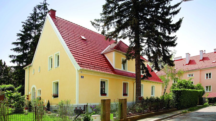 Gelbes Wohnhaus mit rotem Dach und Garten, umgeben von Bäumen und grüner Vegetation in einer Wohngegend.