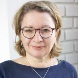 Porträt einer lächelnden Frau mit Brille vor heller Ziegelwand.