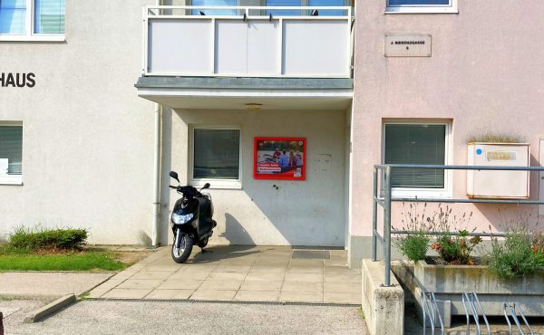 Eingangsbereich mit parkendem Roller und Plakat an einer Hauswand.