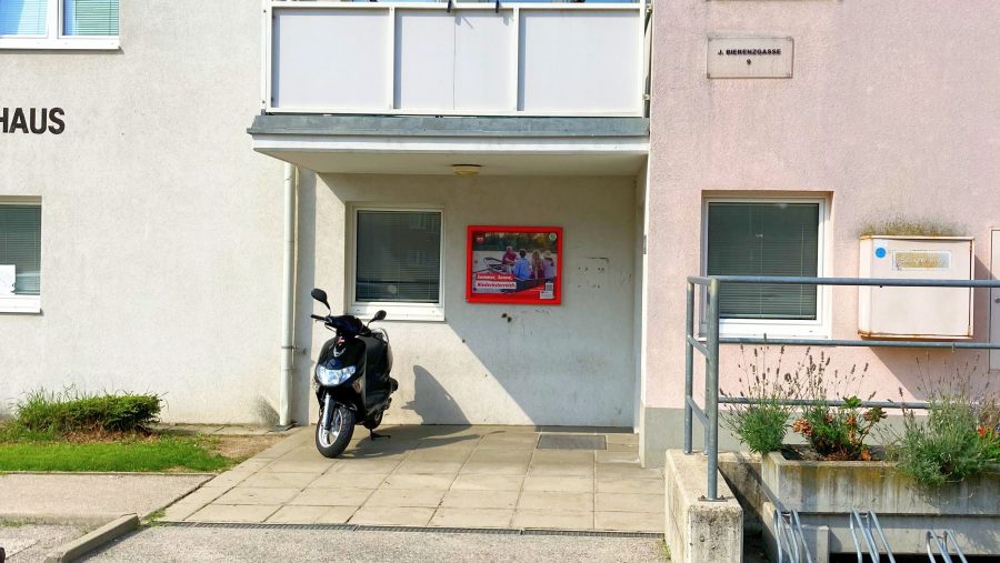 Eingangsbereich mit parkendem Roller und Plakat an einer Hauswand.