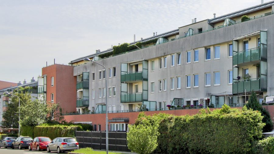 Mehrstöckiges Wohngebäude mit Balkonen und parkenden Autos bei Sonnenschein.