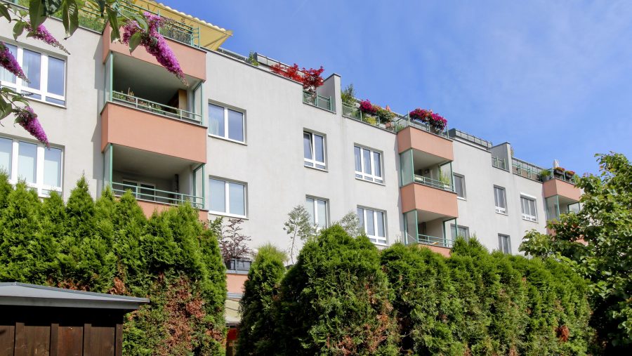 Mehrfamilienhaus mit Balkonen und grünen Bäumen im Vordergrund, sonniger Himmel im Hintergrund.
