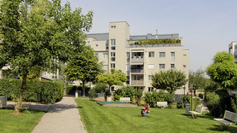 Wohngebäude mit grüner Umgebung, Bäume und Spielplatz im Vordergrund bei Sonnenschein.