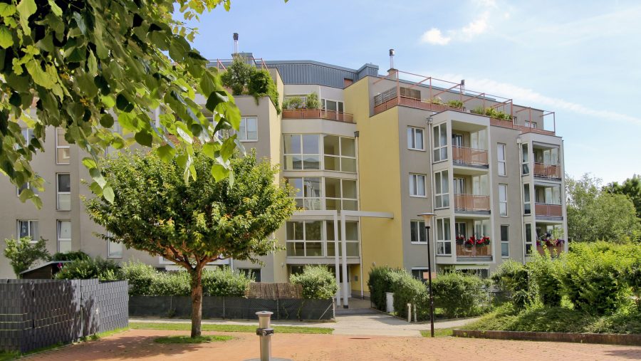 Moderne Wohnanlage mit Bäumen und Garten im Vordergrund, sonniger Tag.