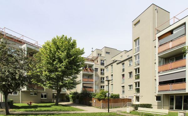 Moderne Wohnanlage mit Bäumen und Grünfläche im Vordergrund an einem sonnigen Tag.