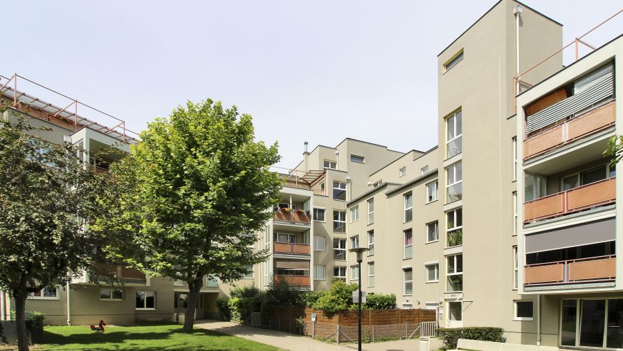 Moderne Wohnanlage mit Bäumen und Grünfläche im Vordergrund an einem sonnigen Tag.