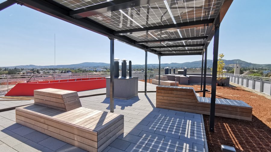 Dachterrasse mit Holzsitzmöbeln und Solardach, Berge im Hintergrund, sonniges Wetter.