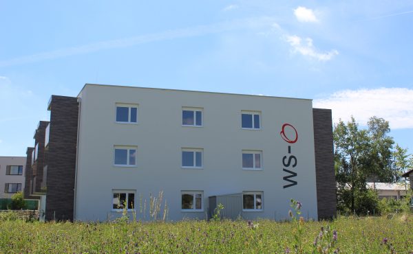 Modernes Mehrfamilienhaus mit Schriftzug, umgeben von Natur. Ideal für Wohnungskauf in ruhiger Lage.