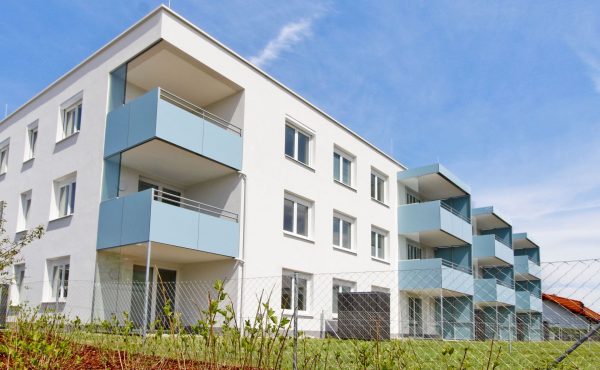 Modernes weißes Mehrfamilienhaus mit blauen Balkonen und grünem Rasen, unter blauem Himmel.
