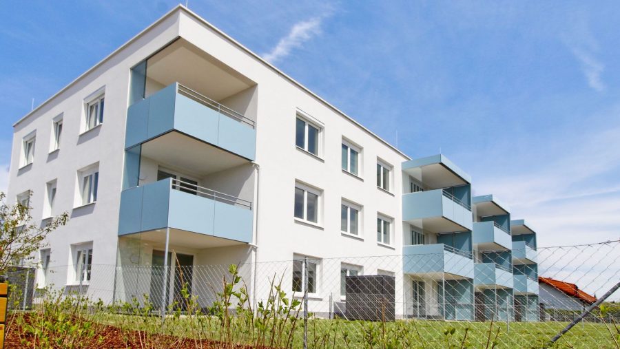 Modernes weißes Mehrfamilienhaus mit blauen Balkonen und grünem Rasen, unter blauem Himmel.