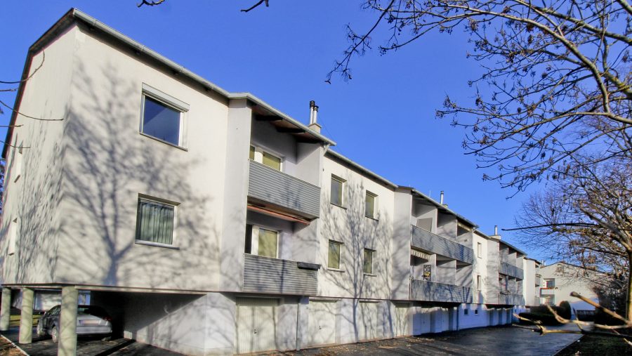 Mehrfamilienhaus an einem sonnigen Tag mit blauem Himmel und kahlen Bäumen im Vordergrund.