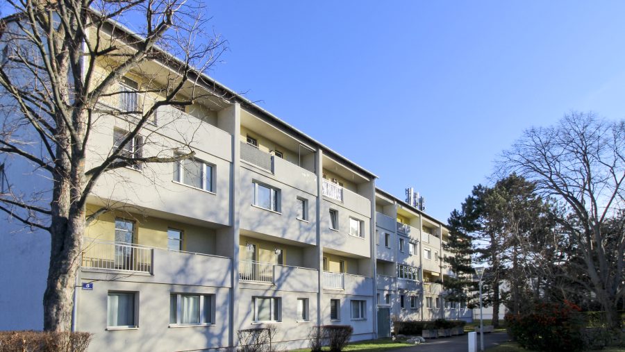 Mehrstöckiges Wohnhaus mit Balkonen an einem sonnigen Tag, umgeben von Bäumen und Gehweg.