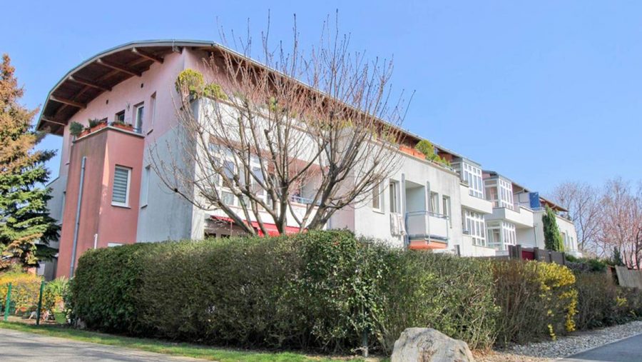 Mehrfamilienhaus mit Garten, Bäumen und Hecken an einem sonnigen Tag.