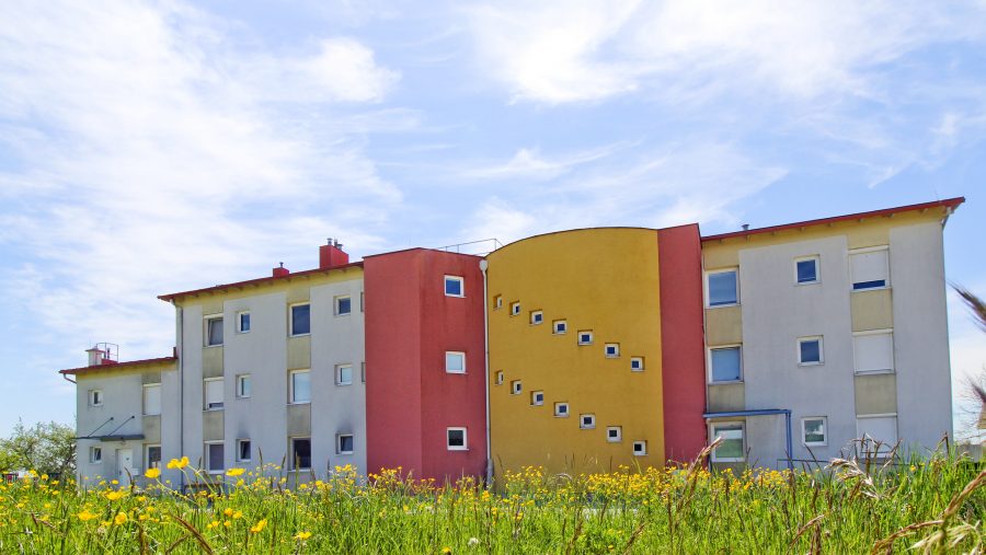 Mehrfamilienhaus mit bunten Fassaden und Blumengarten im Vordergrund unter blauem Himmel.