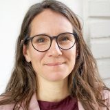 Frau mit Brille lächelt in die Kamera, trägt ein rosa Sakko und ein bordeauxfarbenes Oberteil vor weißer Wand.