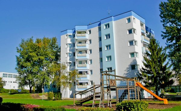 Mehrfamilienhaus mit Spielplatz und Bäumen bei sonnigem Wetter.