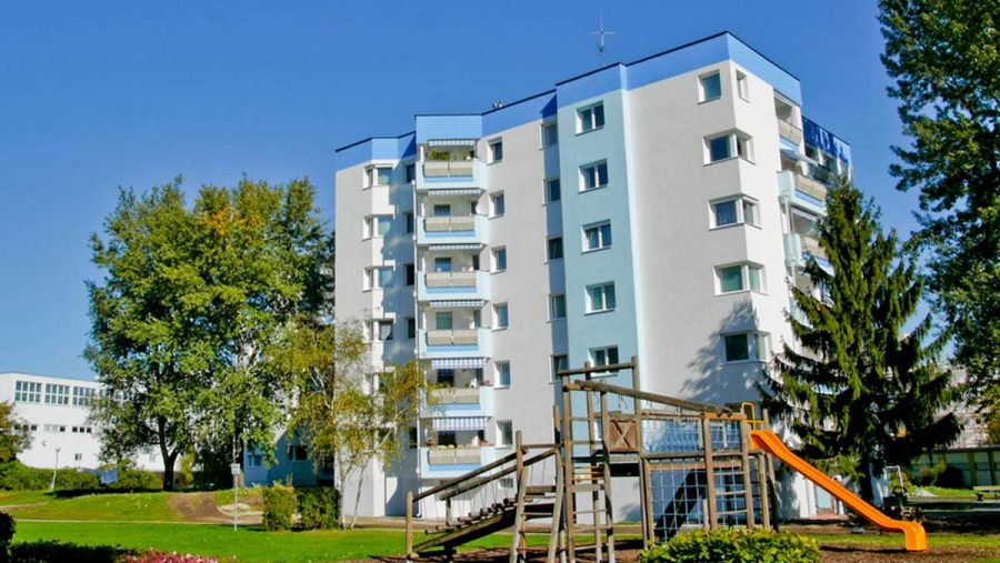 Mehrfamilienhaus mit Spielplatz und Bäumen bei sonnigem Wetter.
