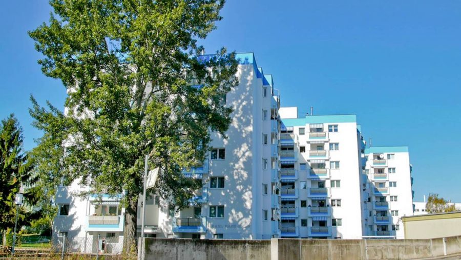 Wohngebäude bei Sonnenschein, umgeben von Bäumen, blauer Himmel im Hintergrund.