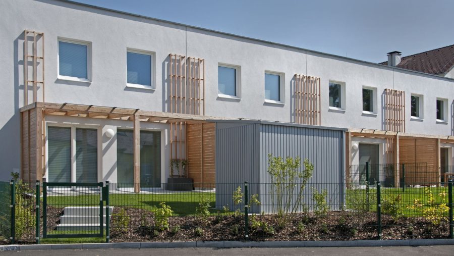 Modernes Reihenhaus mit Garten und Holzelementen, vorne kleiner Garten mit Zaun, blauem Himmel im Hintergrund.