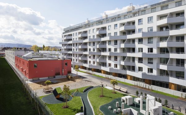 Mehrstöckiger Wohnkomplex mit grünem Spielplatz im Vordergrund bei sonnigem Wetter.
