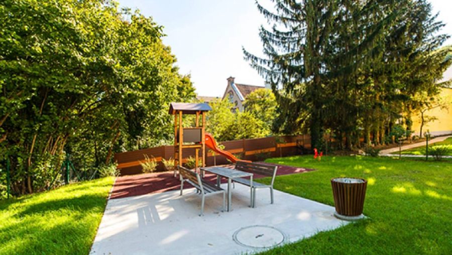 Spielplatz mit Rutsche, Bänken und Bäumen im Hintergrund an einem sonnigen Tag.