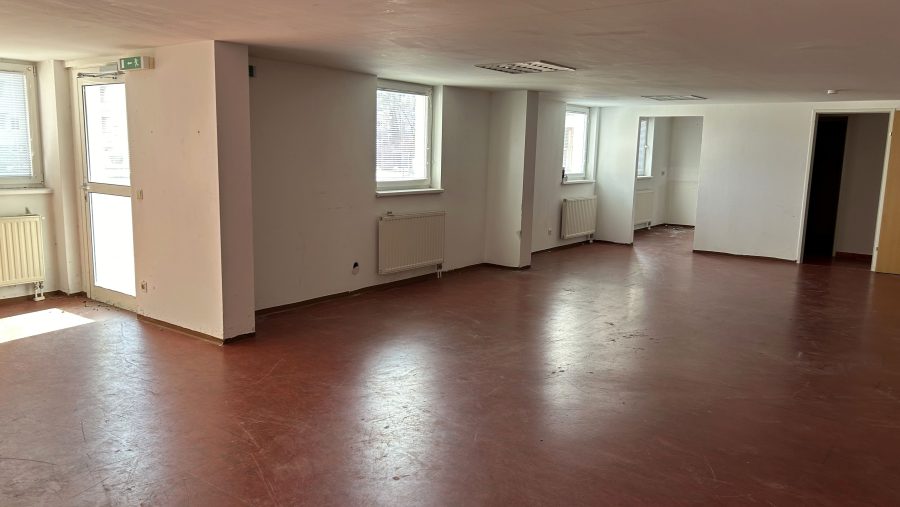 Leerer Raum mit rotem Boden, weißen Wänden, Fenstern und Heizungen, ideal für Büro- oder Wohnnutzung.
