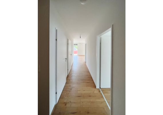 Langer Flur mit Holzboden, weiße Wände und mehreren Türen in einem modernen Gebäude.