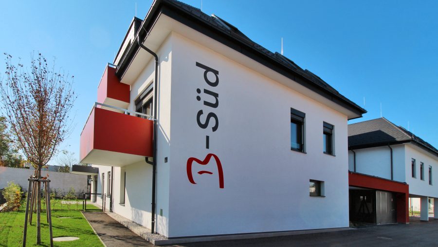 Modernes Gebäude mit weißer Fassade, Balkonen, rotem Logo und klarem Himmel im Hintergrund.