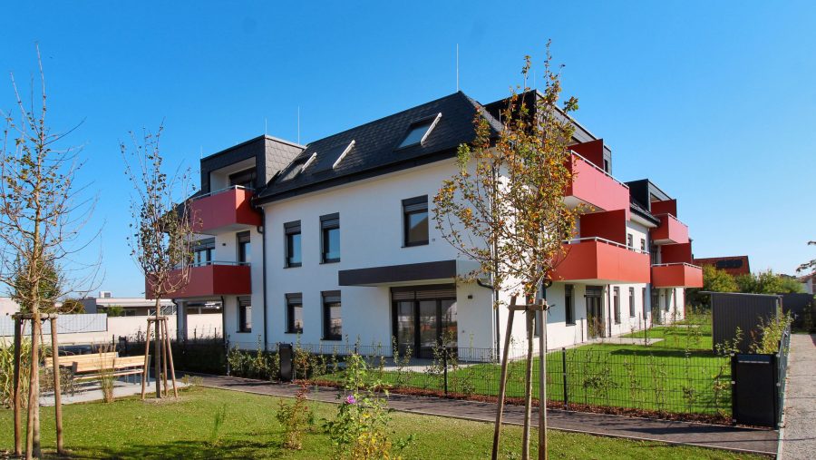 Modernes Wohngebäude mit roten Balkonen an einem sonnigen Tag, umgeben von Bäumen und Rasenflächen.