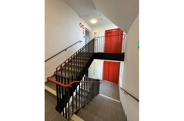 Treppenhaus mit roten Türen und modernen Geländern im Gebäudeobergeschoss.